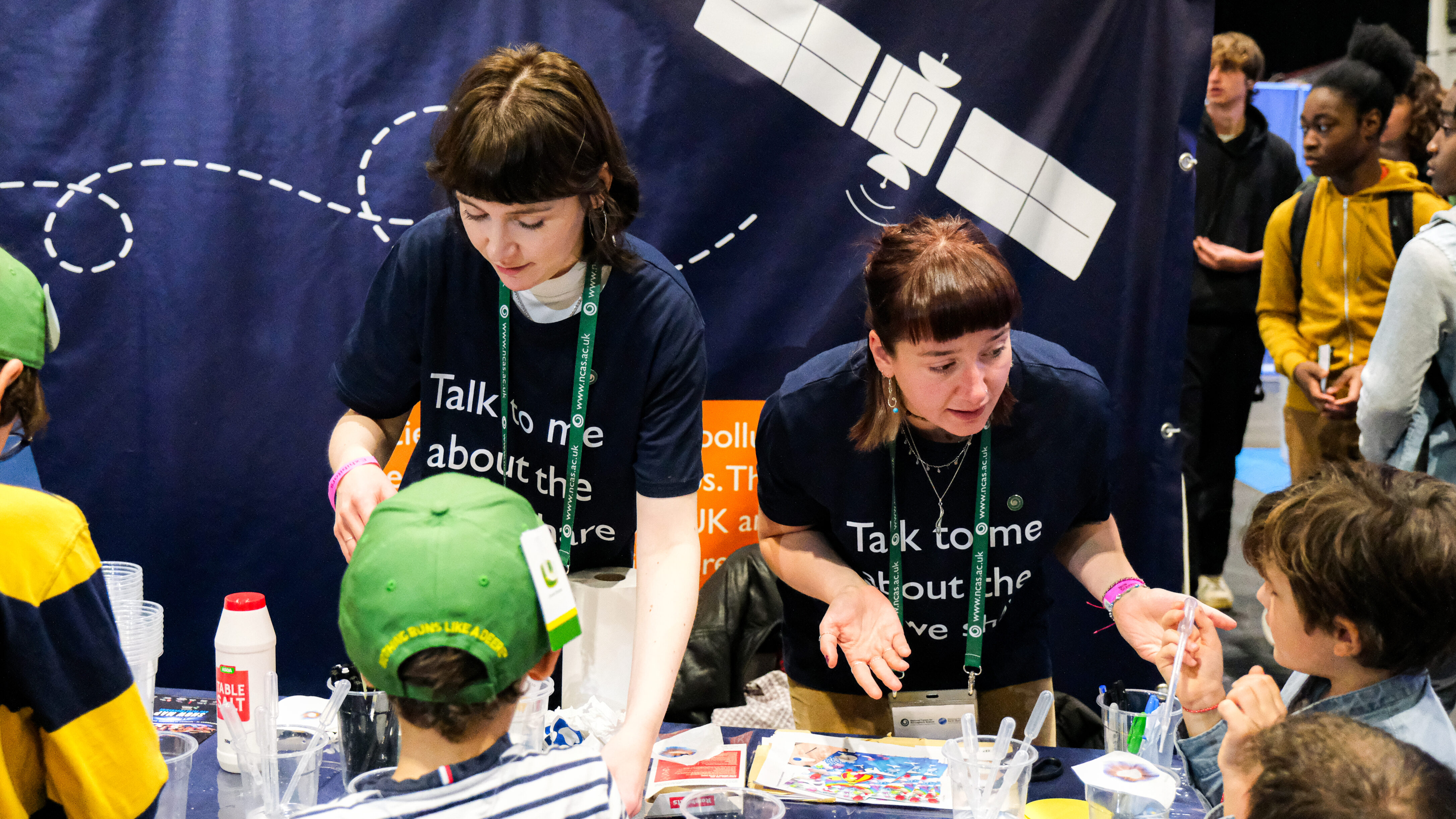 Two people wearing t-shirts that say "Talk to me about the air we share", talking to a group of children. There is a banner featuring a satellite illustration behind them.