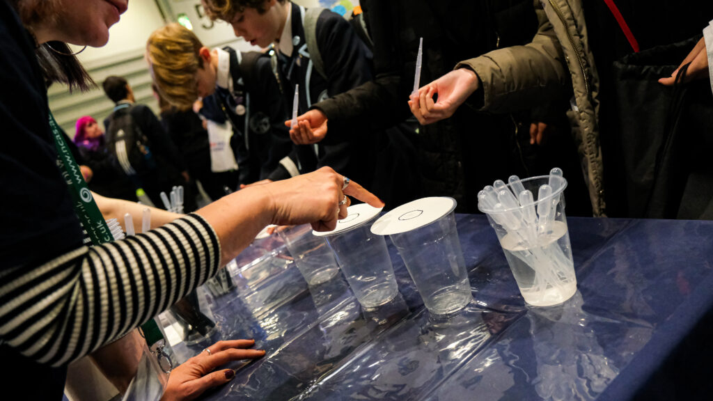 A hand points at a piece of paper which is placed on top of a plastic cup. There is a black circle drawn on the paper. Another hand holds a pipette. More cups, pipettes, and people in the background.