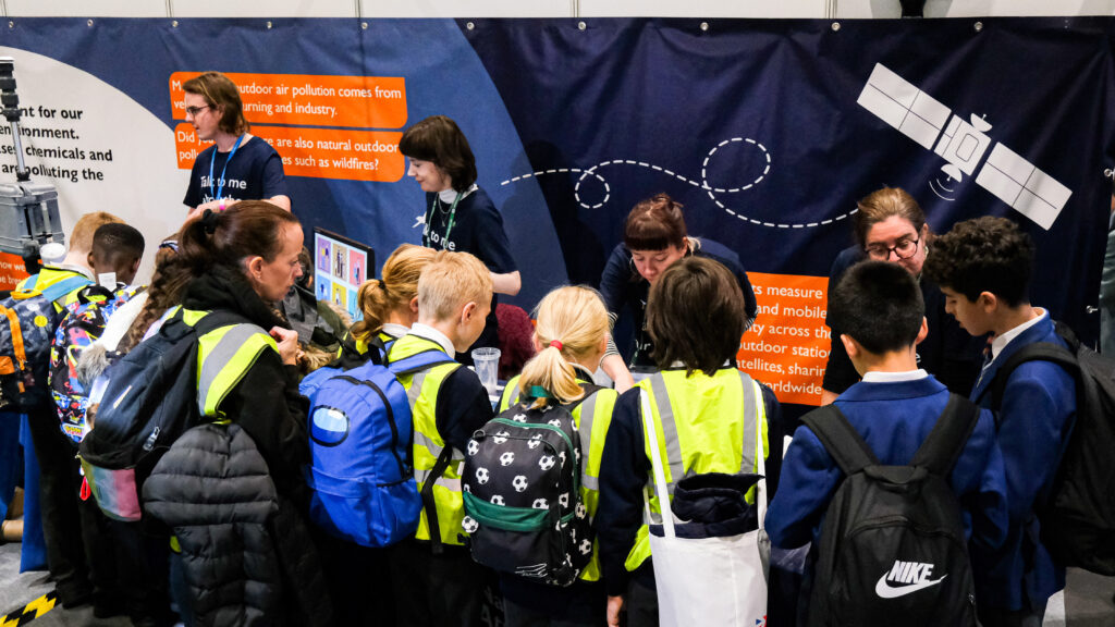 Four people in dark blue t-shirts face a group of children who look at a stand. Behind the stand is a banner featuring a satellite illustration.