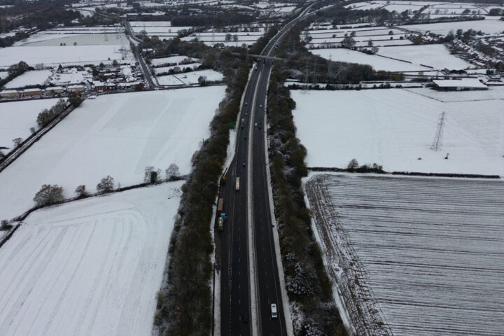 Aerial picture of snow covered fields. Bare trees and hedges line the fields and road travelling through the fields. Cars and lorries are driving on the road.