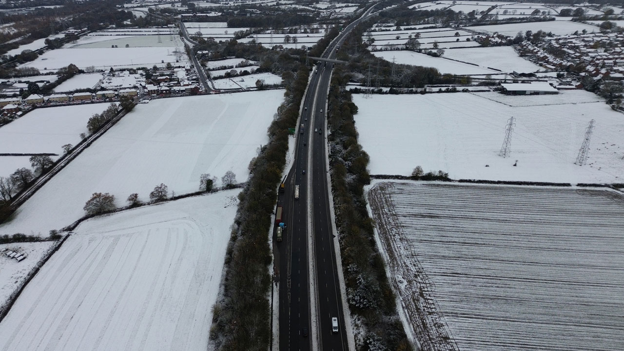 Aerial picture of snow covered fields. Bare trees and hedges line the fields and road travelling through the fields. Cars and lorries are driving on the road.