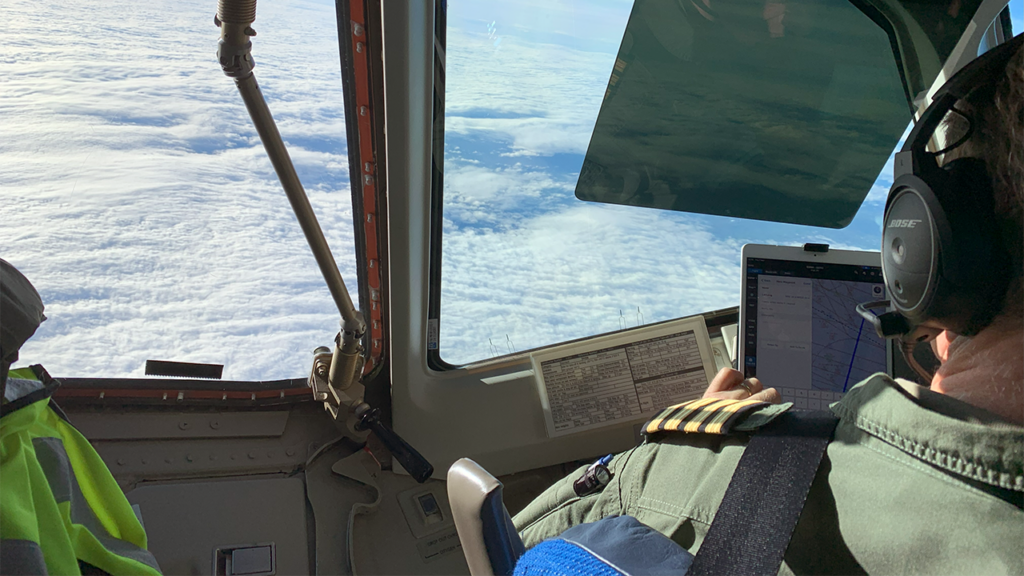 View of a cloudy blue sky from above from within the cockpit of an aircraft. A person looks at a computer display of an aircraft's track - they are wearing headphones and a green uniform.