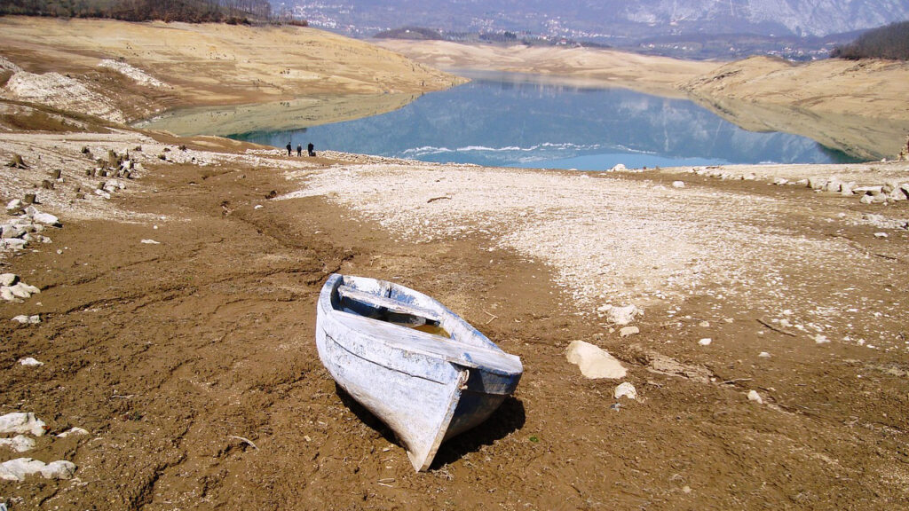 White wooden boat on brown and pale grey dry and stony land, with a blue lake in the background.