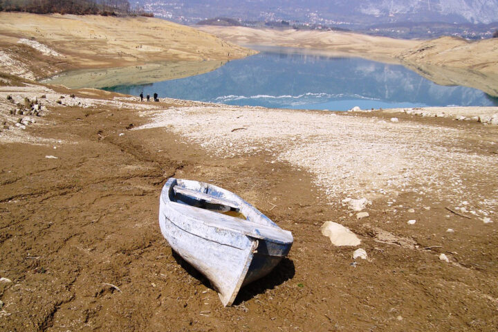 White wooden boat on brown and pale grey dry and stony land, with a blue lake in the background.