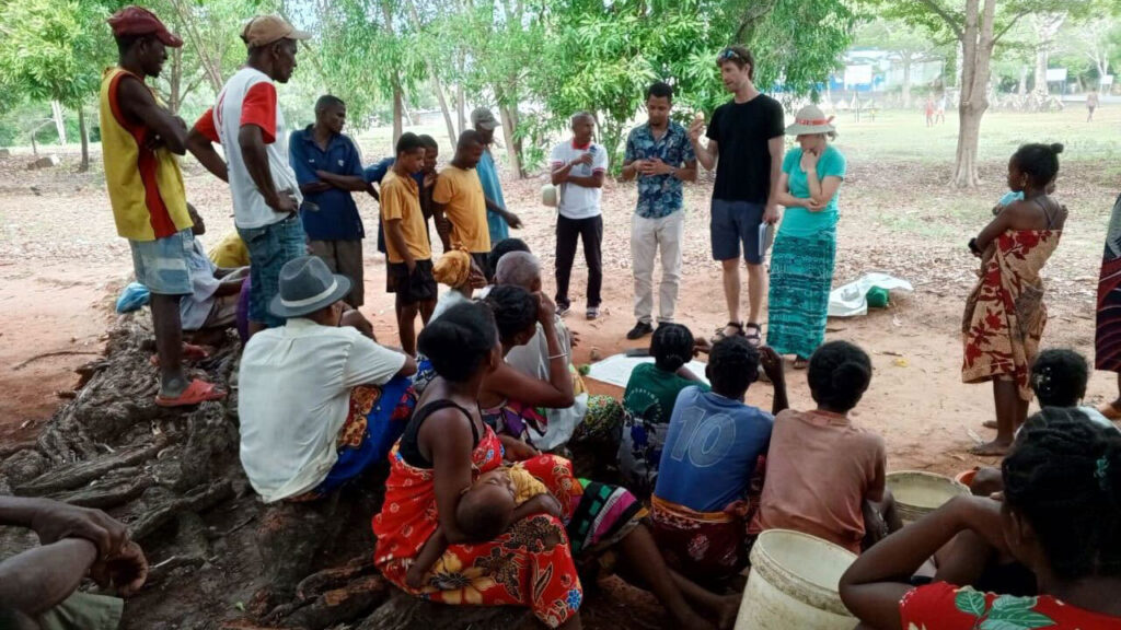 Large group of people sat outside on a tree root. Three people stand at the front of the group and are talking to them. There is a large piece of paper on the floor in front of them.