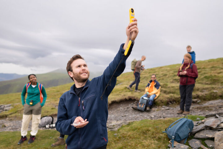 Person wearing a blue fleece holding a yellow weather monitoring instrument in the air. In their other hand is a compass. Green grass, hills and grey clouds in front of them.