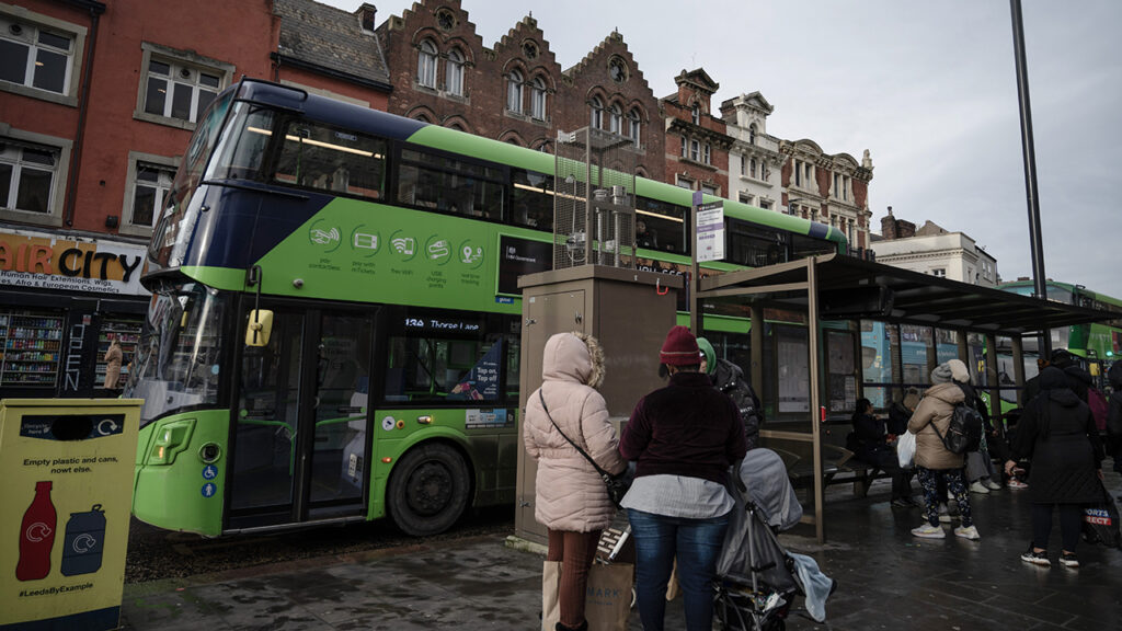 People waiting at a bus stop, with a green double decker bus in the background.