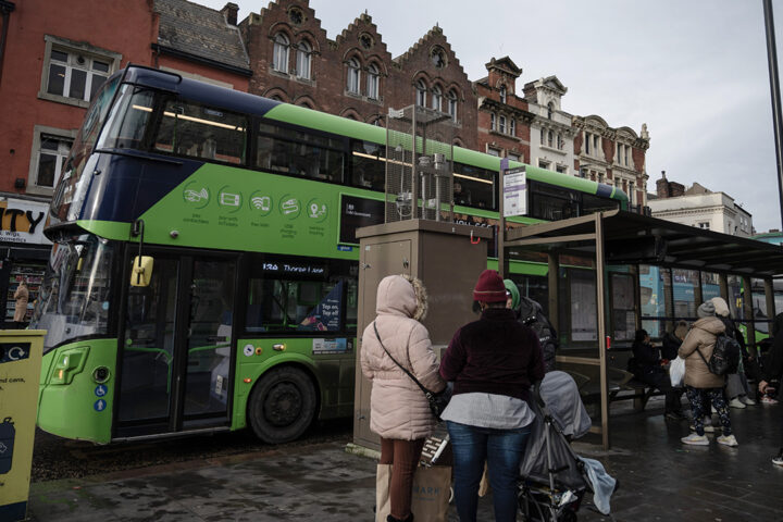 People waiting at a bus stop, with a green double decker bus in the background.