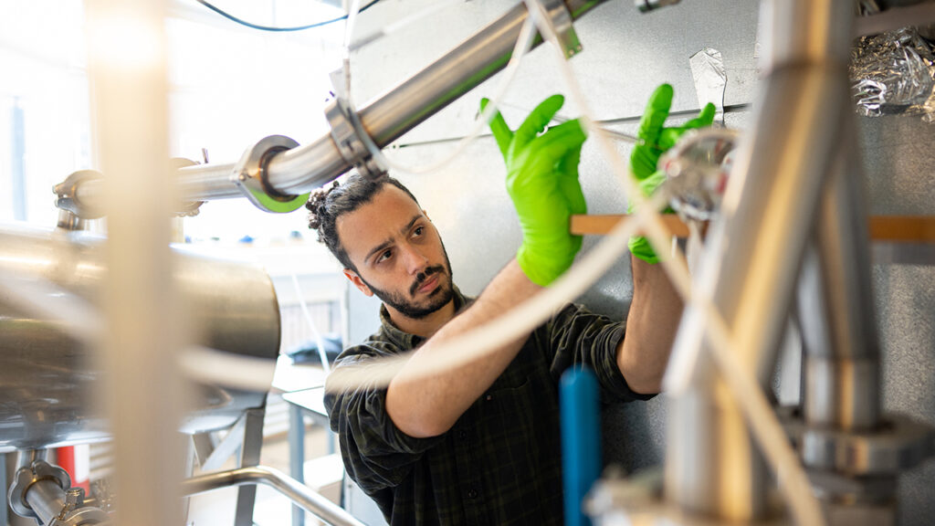 Person with long, tied-up hair and beard wearing green latex gloves holds a syringe next to gas inlet pipes by a large, metal chamber.

