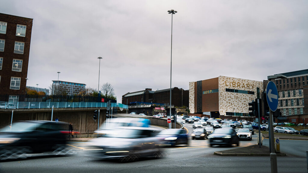 Blurred picture of cars driving in a busy town, with a library building in the background