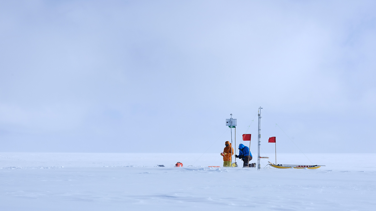 Two people wearing thick outdoor clothing set up a weather mast on snow