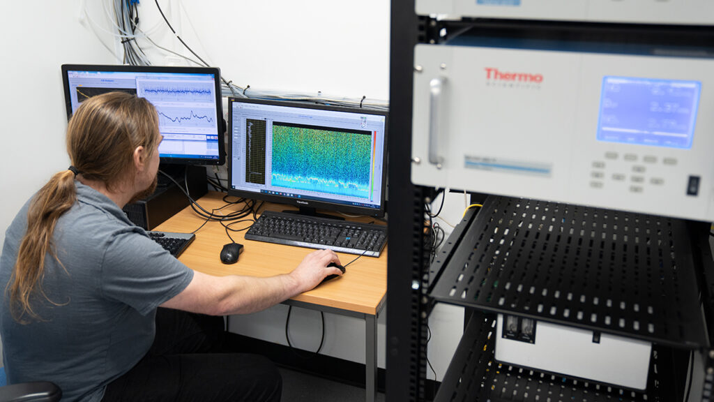 Person with long ponytail and beard sits at a computer desk inside a shipping container filled with racks of scientific equipment, the displays show colourful data visualisations.