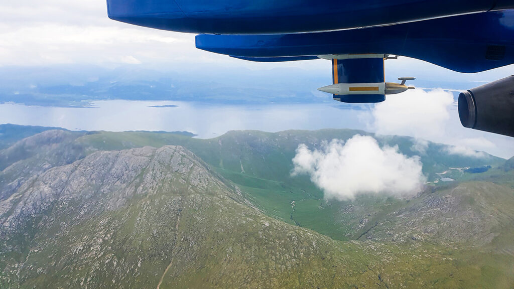 Aerial view of mountains and a lake and fluffy small white clouds from a blue aircraft. Parts of the aircraft are visible in the top right corner of the photo.