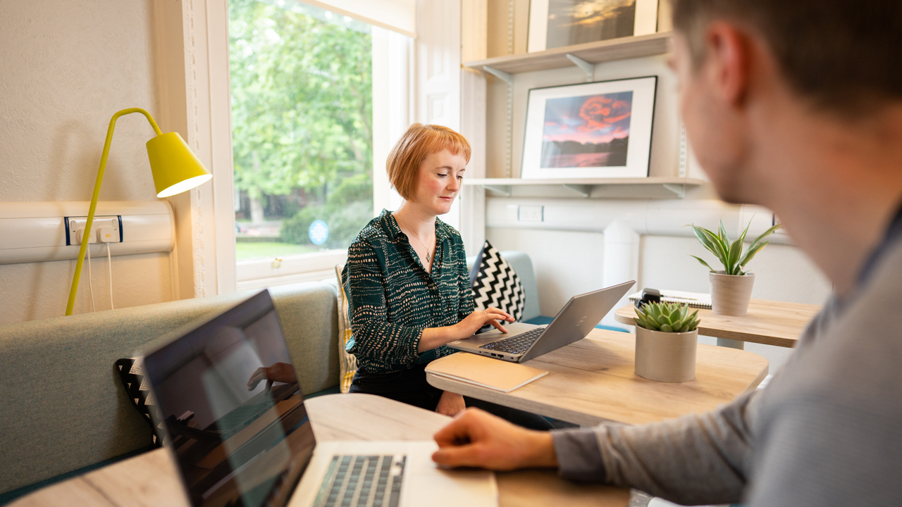 Person wearing green top sits on comfortable chair with a laptop on a small side table, she looks at the screen intently.