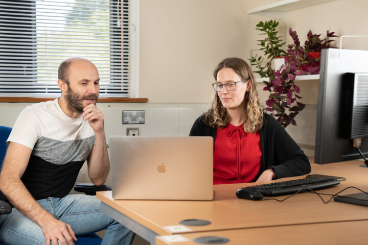 Two people sat at a desk looking at a laptop screen. There is a window in the background, some plants on a shelf on the right hand side, and a computer monitor in the foreground.