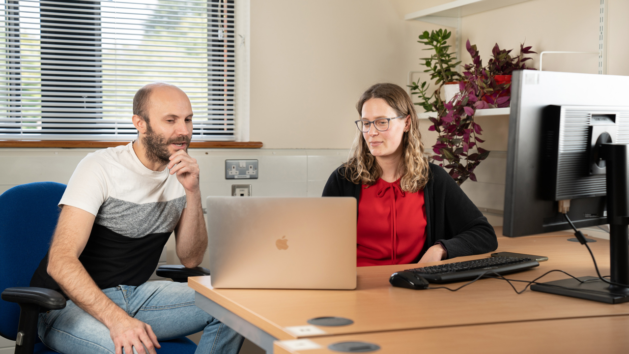 Two people sat at a desk looking at a laptop screen. There is a window in the background, some plants on a shelf on the right hand side, and a computer monitor in the foreground.