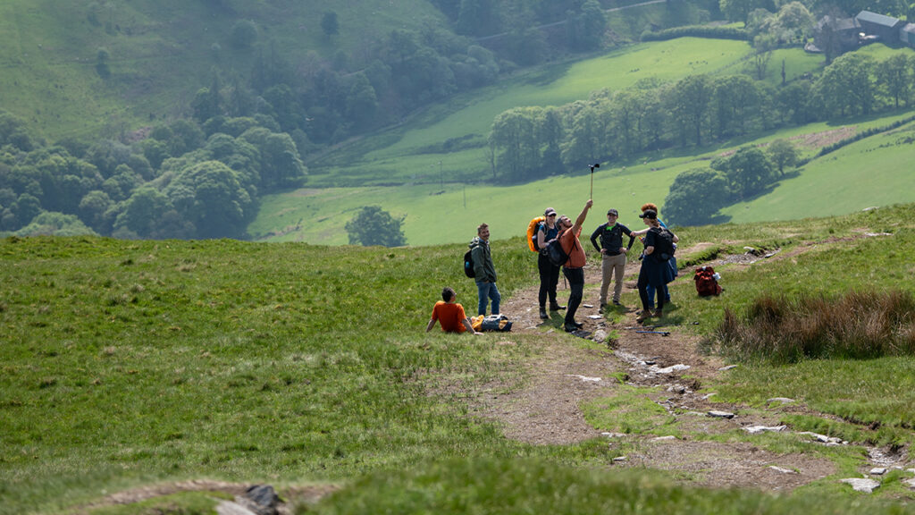 Group of seven people stood on a green hill. Green valley with hills, tree, houses and a lake in the background.