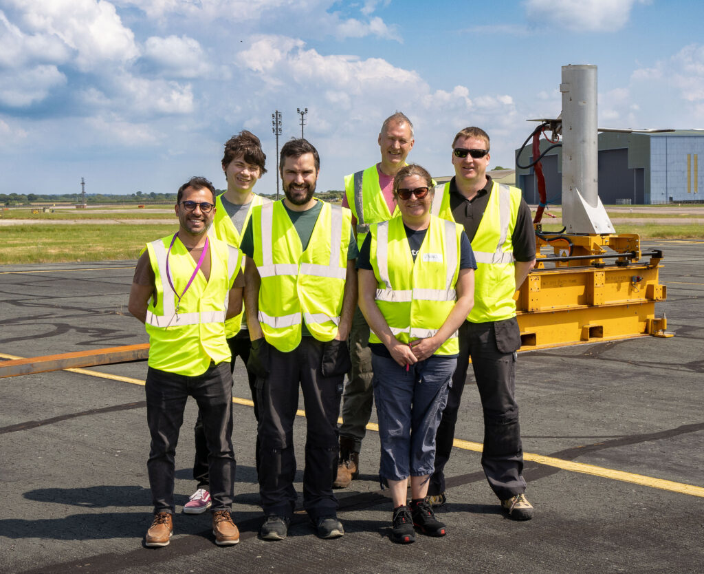 Six people in hi-vis jackets in front of a large piece of emissions measuring equipment at an airport. They are smiling at the camera.