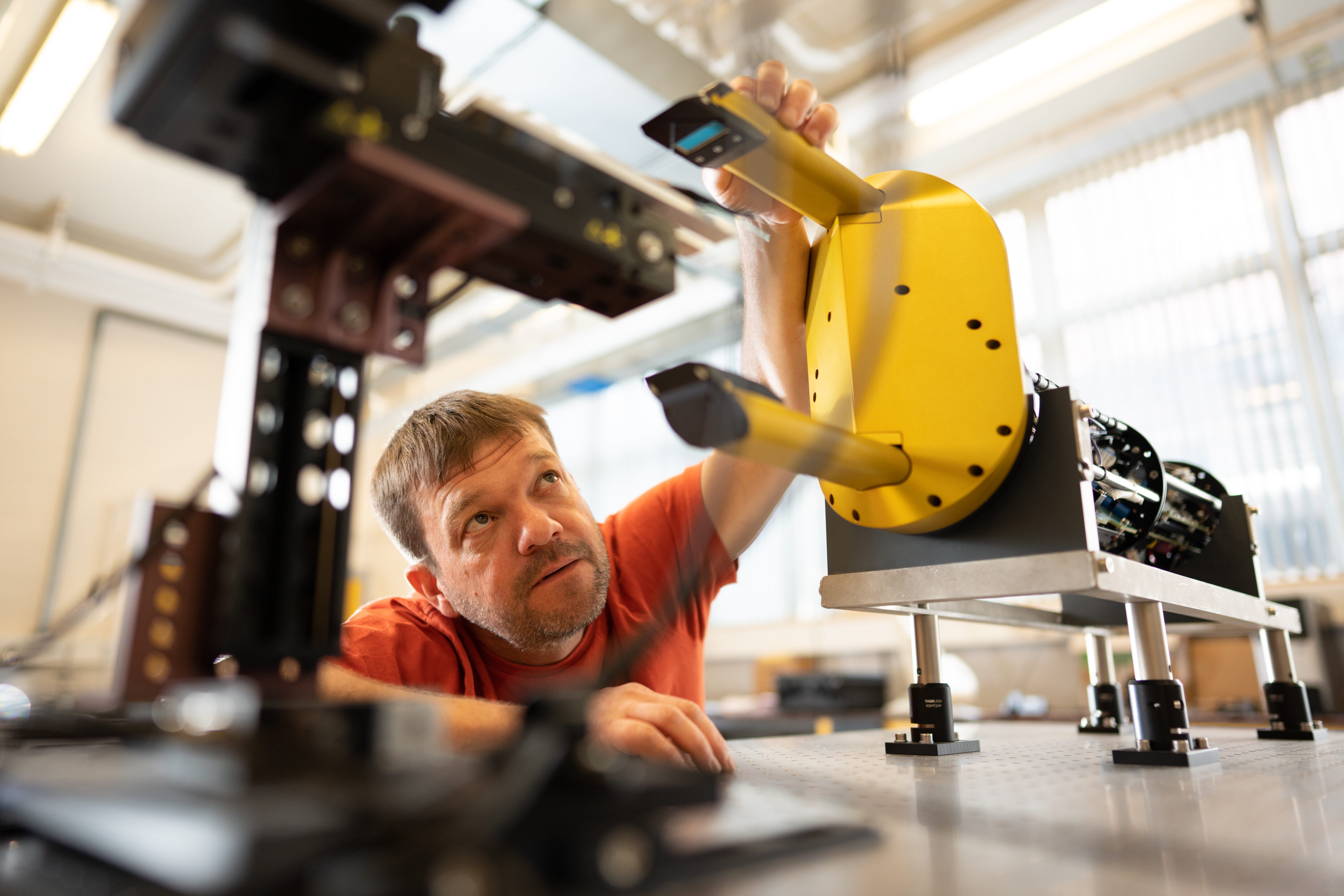 Person with short hair wearing orange top sits in laboratory space. They reach up towards a large yellow scientific instrument, where two opposing measurement probes face each other.