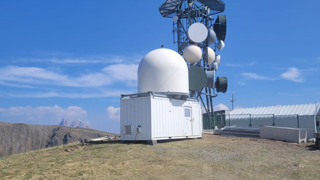 White radar dome on top of a white shipping container, mounted on a grassy hill, with mountains and a blue sky in the background