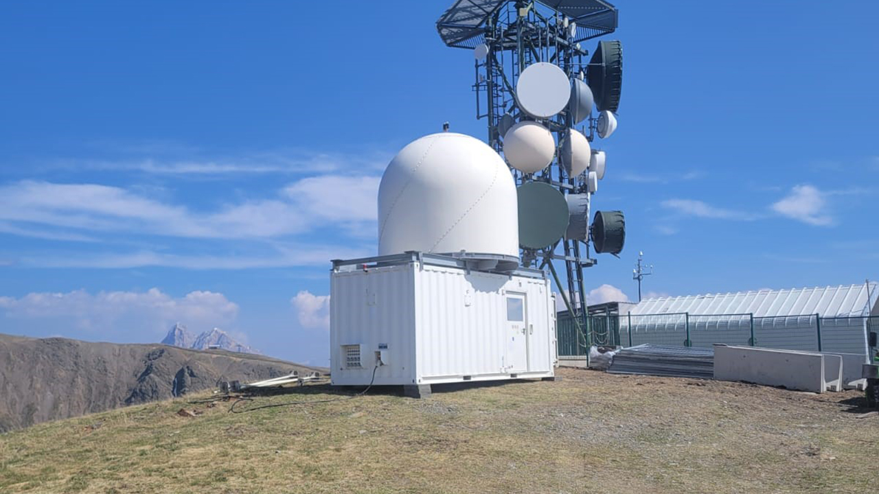 White radar dome on top of a white shipping container, mounted on a grassy hill, with mountains and a blue sky in the background