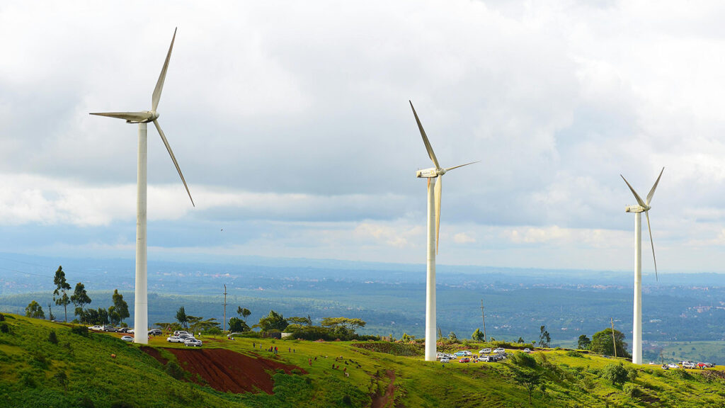 3 white wind turbines on a grassy green slope, with people walking underneath, and a grey-blue cloudy sky in the background.