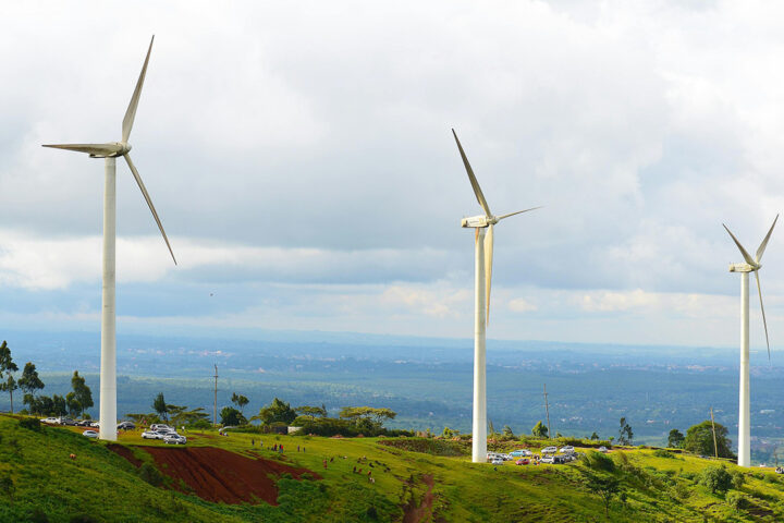 3 white wind turbines on a grassy green slope, with people walking underneath, and a grey-blue cloudy sky in the background.