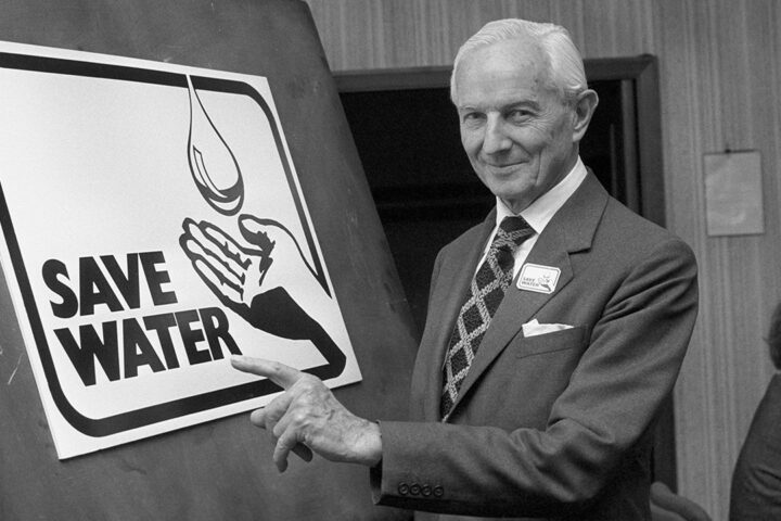 Black and white photo of a man wearing a suit and tie, standing next to a poster that says save water.