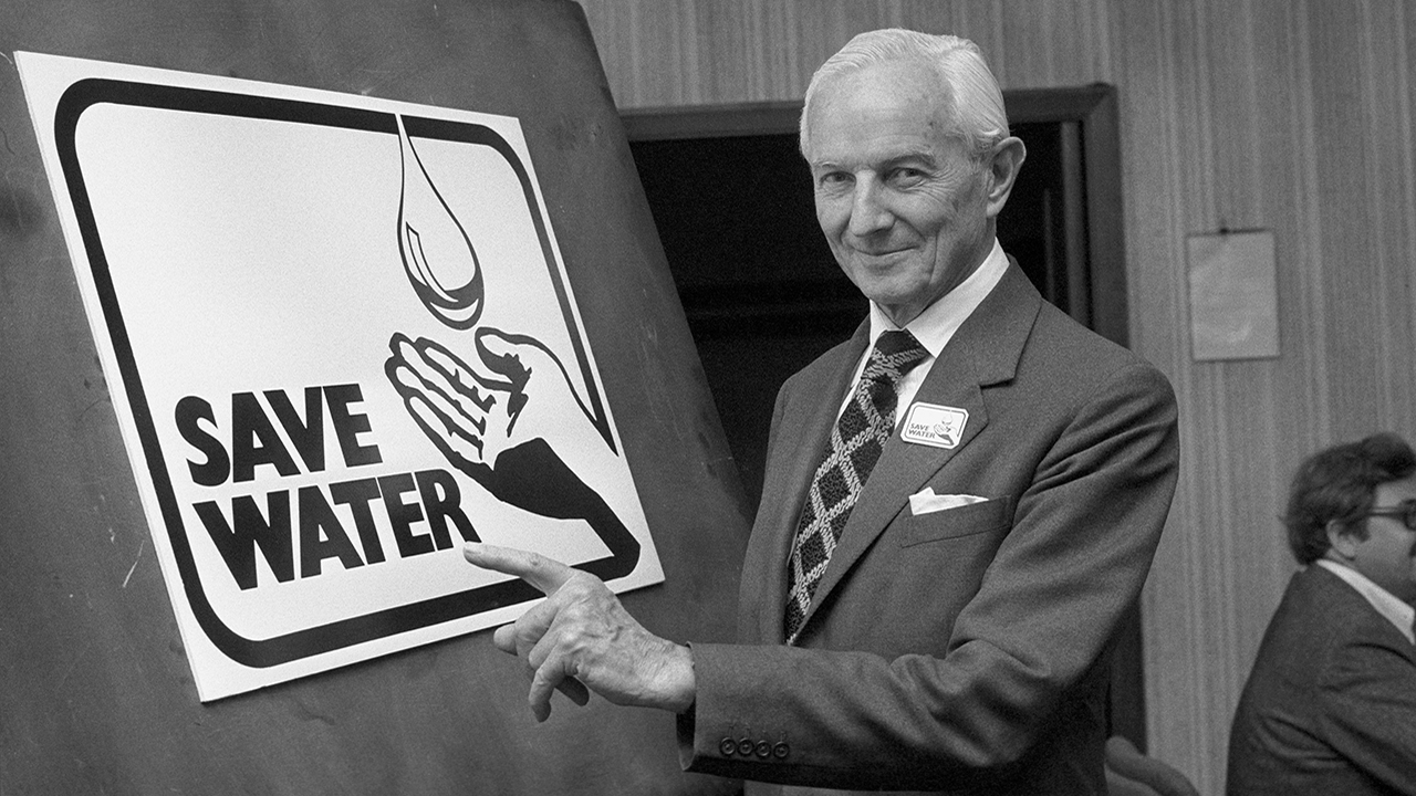 Black and white photo of a man wearing a suit and tie, standing next to a poster that says save water.