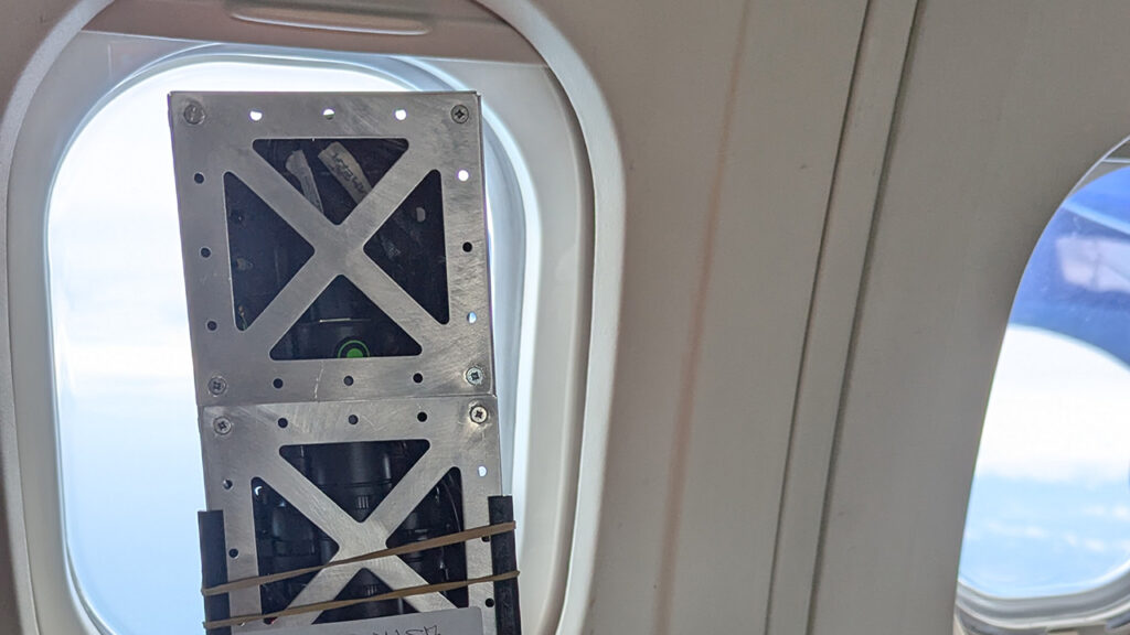 A small satellite next to the inside of an aircraft window, looking out onto blue sky.