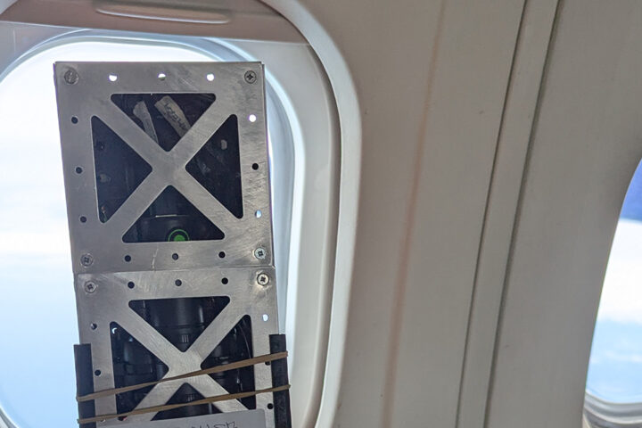 A small satellite next to the inside of an aircraft window, looking out onto blue sky.