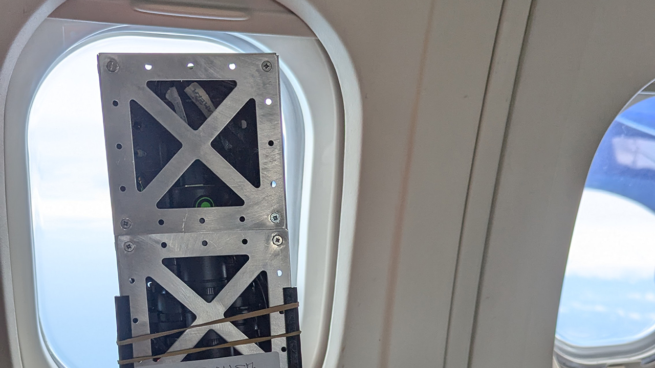 A small satellite next to the inside of an aircraft window, looking out onto blue sky.