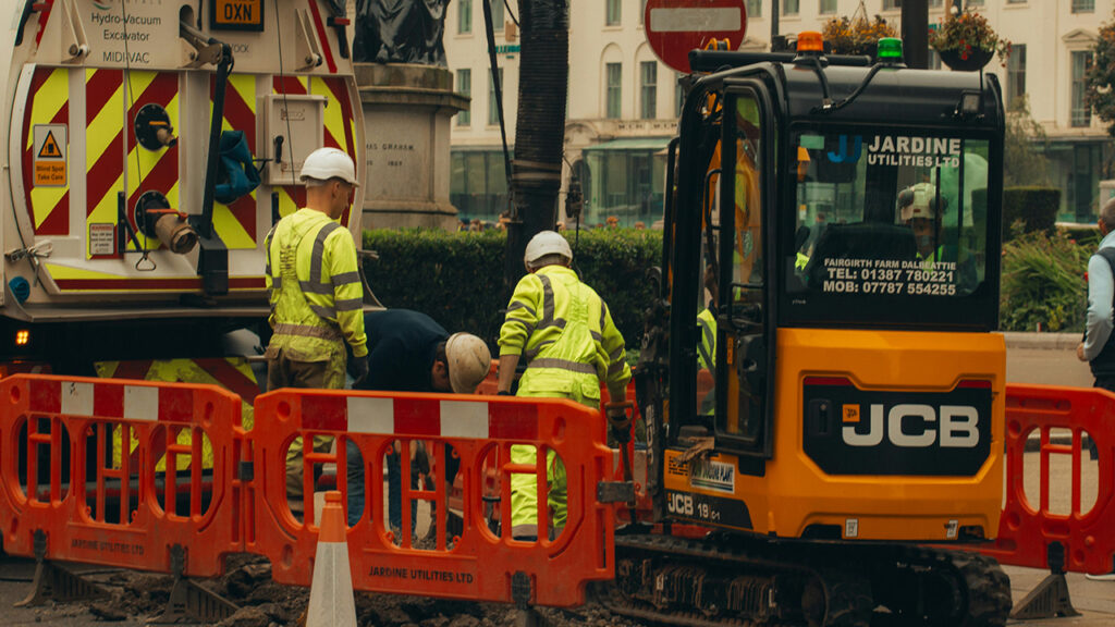 Two construction workers wearing hi vis jackets stand behind bright orange barriers, next to a small JCB digger