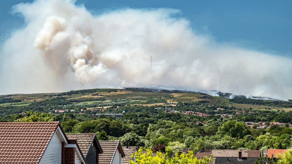 Big white smoky cloud rises up on a green hill, with a blue sky in the background, and people's houses in the foreground.
