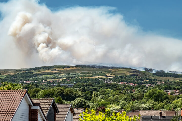 Big white smoky cloud rises up on a green hill, with a blue sky in the background, and people's houses in the foreground.