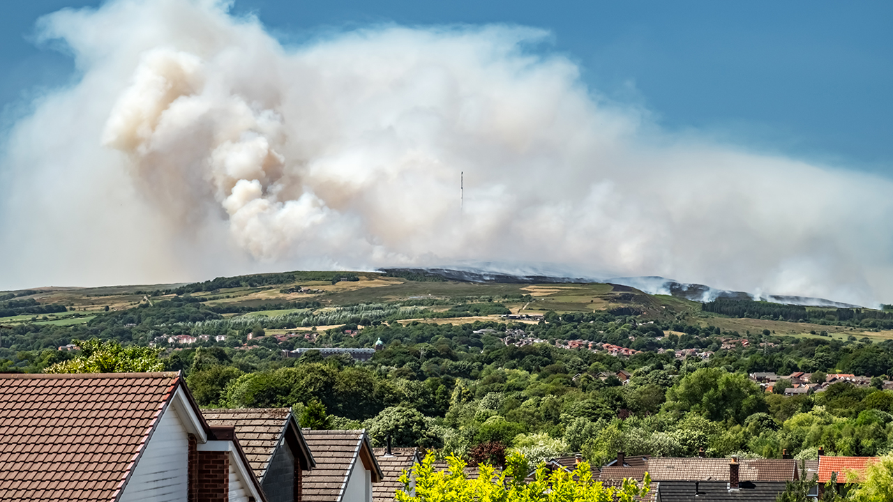 Big white smoky cloud rises up on a green hill, with a blue sky in the background, and people's houses in the foreground.