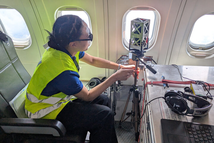 A person wearing a hi vis jacket sits in a seat on an aircraft, reaching towards a small satellite that is positioned next to an aircraft window