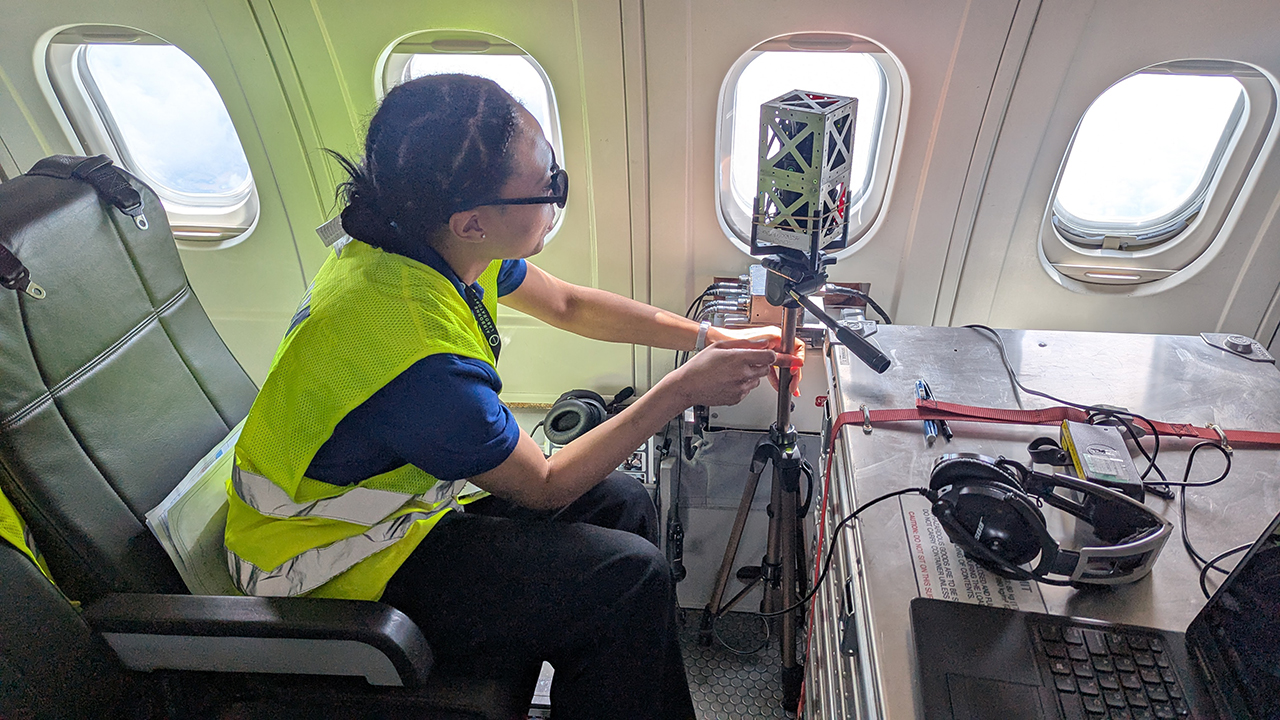 A person wearing a hi vis jacket sits in a seat on an aircraft, reaching towards a small satellite that is positioned next to an aircraft window