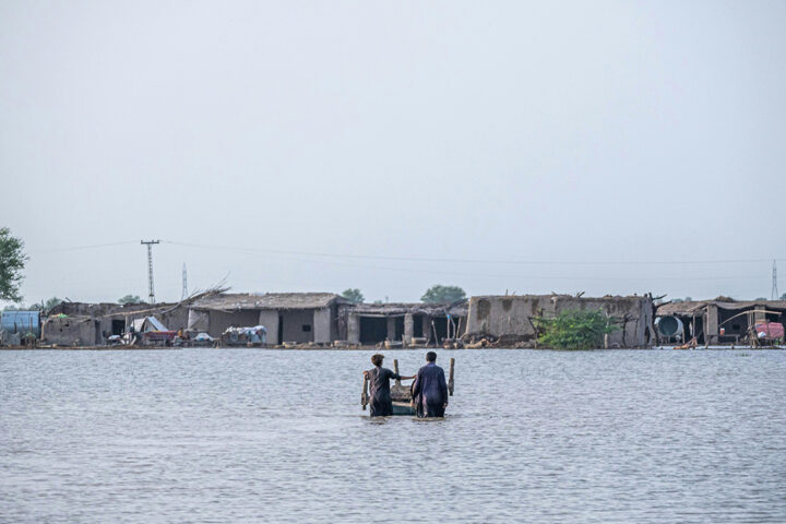 Two people stand wait deep in floodwater, in front of some small buildings and trees