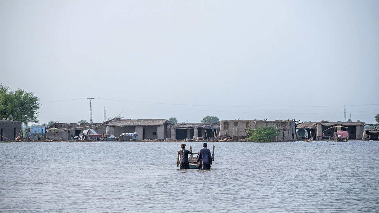Two people stand wait deep in floodwater, in front of some small buildings and trees