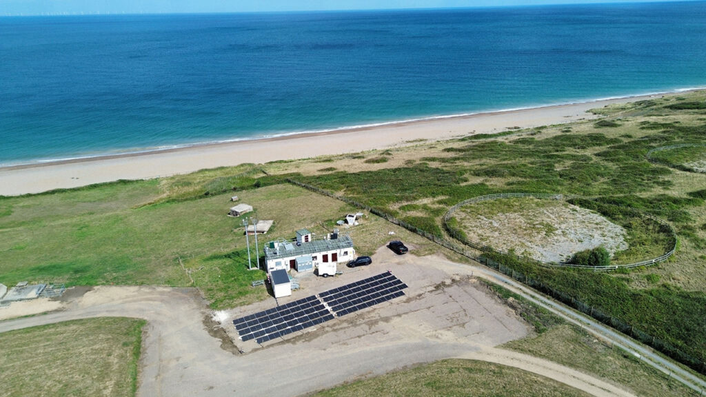 Array of solar panels on a small car park near some buildings, surrounded by green grass and next to a sandy beach and blue sea.