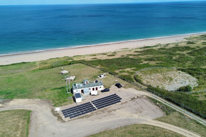 Array of solar panels on a small car park near some buildings, surrounded by green grass and next to a sandy beach and blue sea.