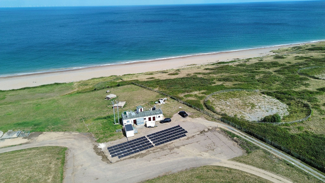 Array of solar panels on a small car park near some buildings, surrounded by green grass and next to a sandy beach and blue sea.