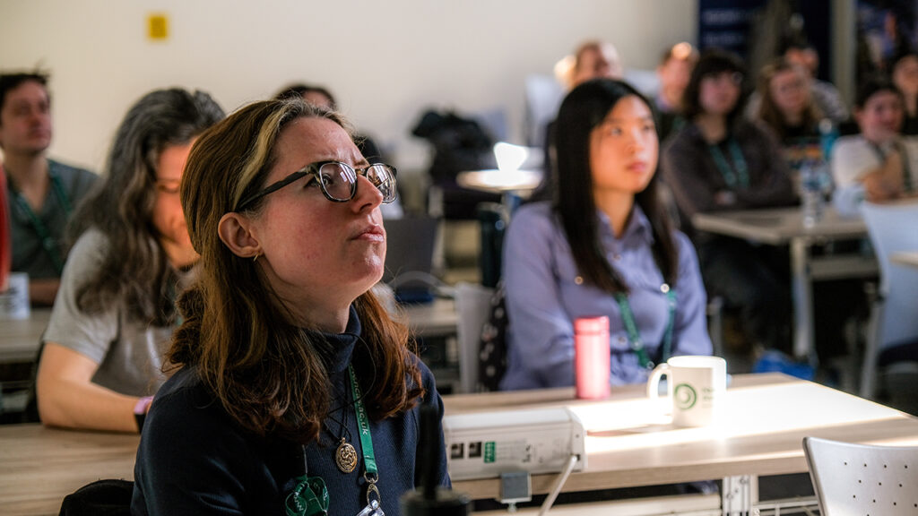 Person wearing glasses, with shoulder length hair, with people sat at desks in the background.