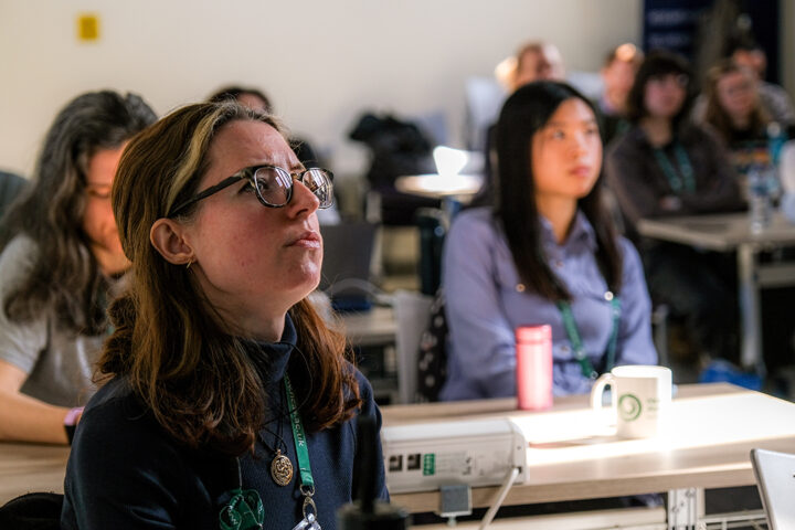 Person wearing glasses, with shoulder length hair, with people sat at desks in the background.