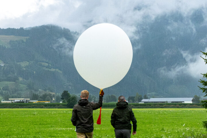 Two people stood in a green field, looking towards a hillside. One of the people is holding a large white balloon.