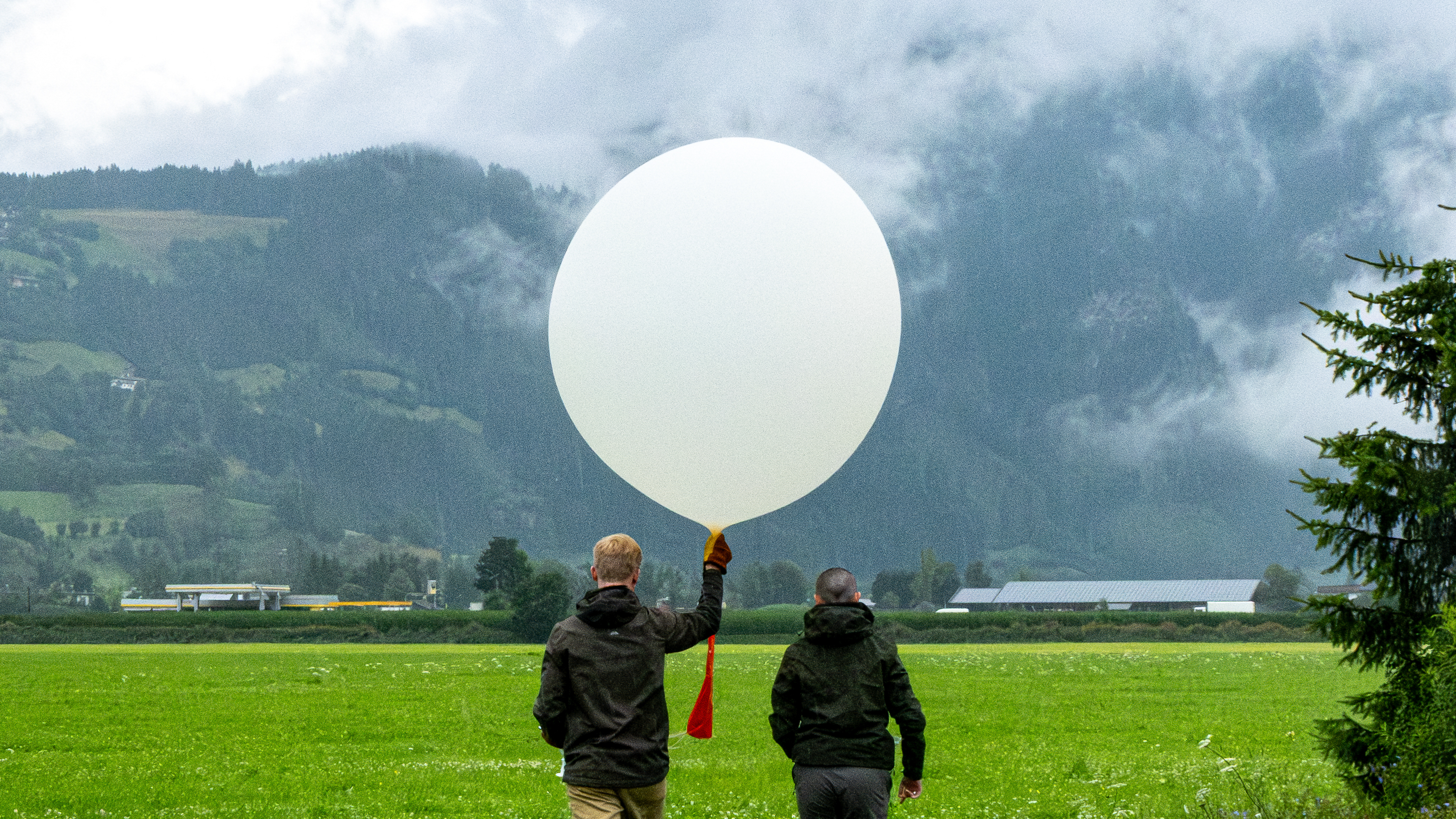 Two people stood in a green field, looking towards a hillside. One of the people is holding a large white balloon.