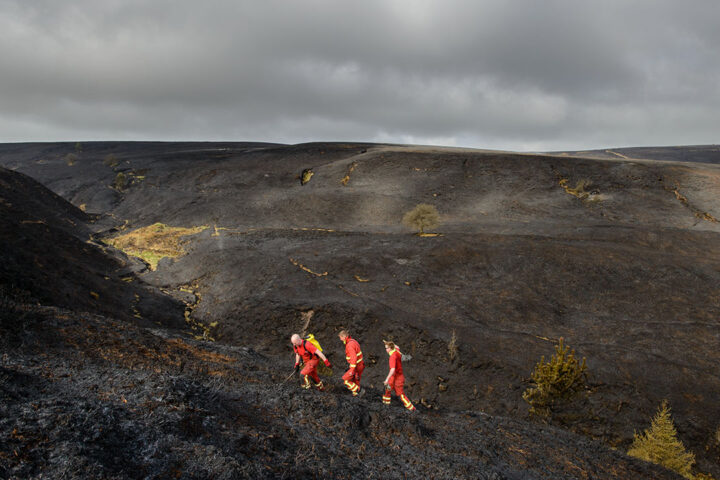 Black burnt moorland landscape, still smouldering, with grey cloudy skies, and 3 firefighters wearing orange uniforms walking uphill from the right