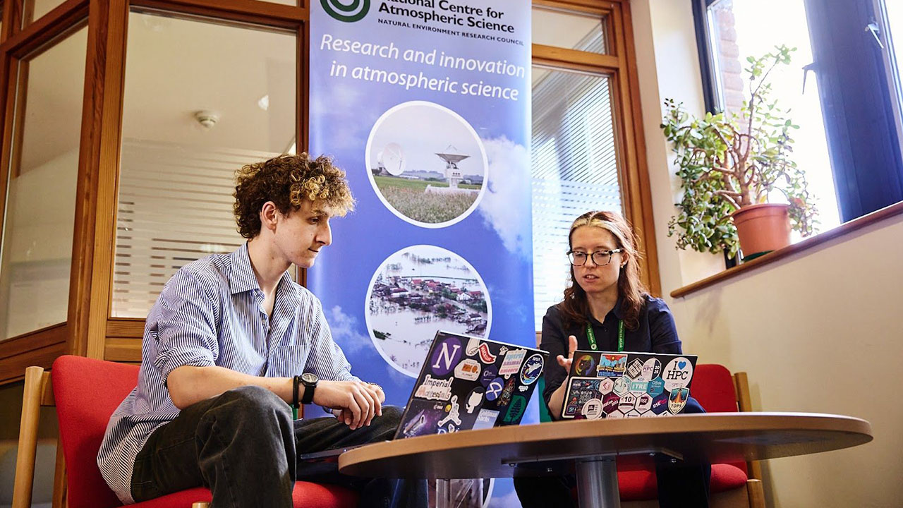 Two people sat on red chairs at a small round low table, looking at their laptops.