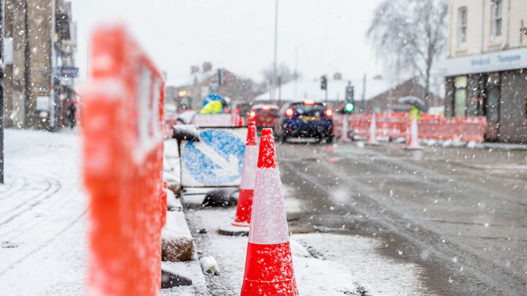 Traffic cones and construction barriers on a snowy street, with cars in the distance driving between roadworks